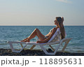 A young woman relaxes on a beach lounge chair by the ocean, sipping a refreshing drink under the warm sun. The serene coastal scenery and clear blue water capture the essence of a perfect summer day. 118895631