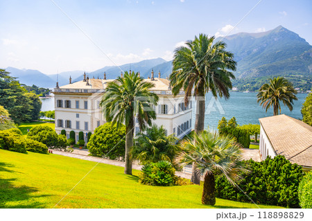 Nestled by the tranquil waters of Lake Como, Villa Melzi showcases neoclassical architecture amidst lush gardens and majestic mountains under a bright blue sky. 118898829