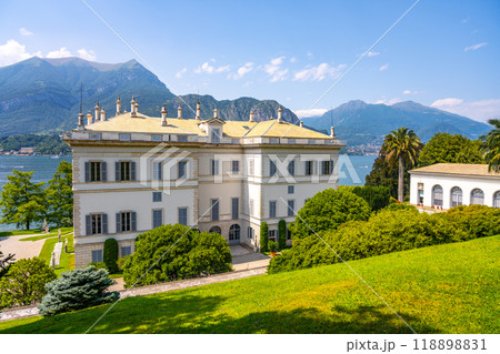 The stunning Villa Melzi stands elegantly by Lake Como, surrounded by lush gardens and majestic mountains under a clear blue sky. Its neoclassical design showcases intricate details and beauty. 118898831