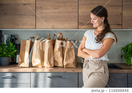 Young woman next to paper bags full of food in kitchen. Groceries ordered online and delivered by courier Young woman next to paper bags full of food in kitchen. Groceries ordered online and delivered by courier 118898848