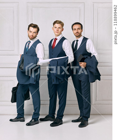 Discussing business after productive day. Young men dressed formal attire posing, against white background with blazers in hands. Discussing business after productive day. Young men dressed formal attire posing, against white background with blazers in hands. 118899462