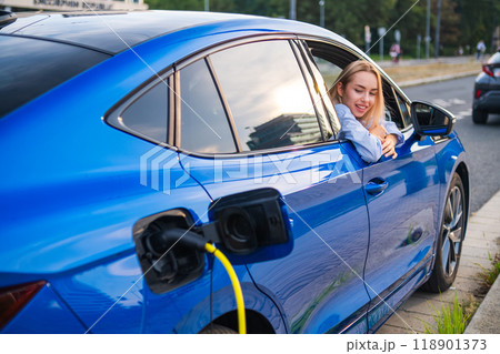 Blonde woman relaxes in her electric vehicle while it charges at a city charging station. 118901373