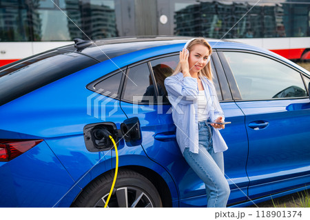 Woman, listening to music through headphones, uses her phone to check the charging progress of her electric car. Woman, listening to music through headphones, uses her phone to check the charging progress of her electric car. 118901374