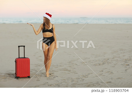 Happy young woman in red Christmas Santa Claus hat and bikini with suitcase on sand seashore shows hand for advertising booking online ticket, hot tours for travels New Year holidays.Winter vacations. Happy young woman in red Christmas Santa Claus hat and bikini with suitcase on sand seashore shows hand for advertising booking online ticket, hot tours for travels New Year holidays.Winter vacations. 118903704