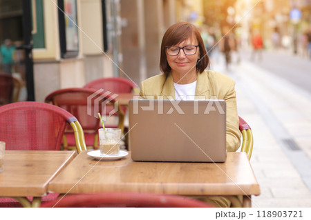 Mature stylish businesswoman in eyeglasses works on laptop while sitting with a coffee drink at European cafe outdoors. Concept of remote work from public place, digital freelance and modern lifestyle 118903721