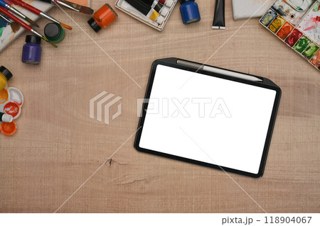 Digital tablet with empty screen and variety of art supplies on a wooden table. Top view 118904067