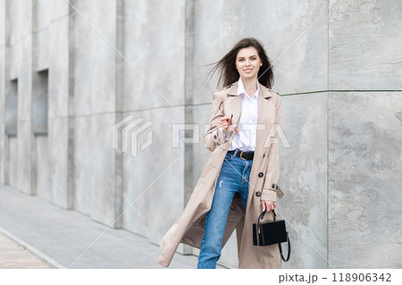 A young pretty girl in a raincoat and jeans walks around the city near a tall building, with a handbag in hand A young pretty girl in a raincoat and jeans walks around the city near a tall building, with a handbag in hand 118906342