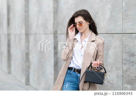 A young pretty girl in a raincoat and jeans walks around the city near a tall building, with a handbag in hand 118906343