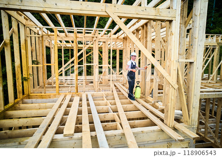 Laborer constructing wooden frame house near forest. Man treating woods, applying fire retardant using sprayer, while dressed in protective suit, helmet. Concept of modern eco-friendly construction. Laborer constructing wooden frame house near forest. Man treating woods, applying fire retardant using sprayer, while dressed in protective suit, helmet. Concept of modern eco-friendly construction. 118906543