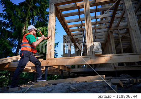 Carpenter constructing two-story wooden frame house near the forest. Bearded man hammering nails while wearing protective helmet and construction vest. Concept of modern ecological construction. Carpenter constructing two-story wooden frame house near the forest. Bearded man hammering nails while wearing protective helmet and construction vest. Concept of modern ecological construction. 118906544