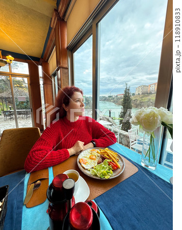 Millennial woman in a red sweater sits at a restaurant table by the window. In window scenic river view enhances the calm and reflective mood. Breakfast in hotel cafe and good start of the new day Millennial woman in a red sweater sits at a restaurant table by the window. In window scenic river view enhances the calm and reflective mood. Breakfast in hotel cafe and good start of the new day 118910833