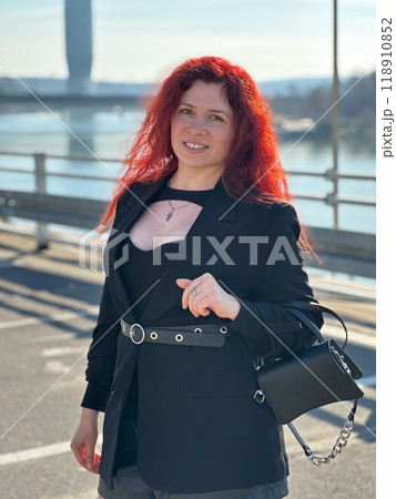 Millennial woman with long curly red hair gazing thoughtfully at the camera on a sunny day. Background features modern buildings and a river, evoking a sense of urban calm. City life, Belgrade and Millennial woman with long curly red hair gazing thoughtfully at the camera on a sunny day. Background features modern buildings and a river, evoking a sense of urban calm. City life, Belgrade and 118910852