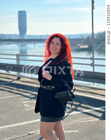 Millennial woman with long curly red hair gazing thoughtfully at the camera on a sunny day. Background features modern buildings and a river, evoking a sense of urban calm. City life, Belgrade and 118910854