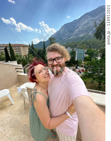 Millennial couple enjoys a vacation with a beautiful mountain backdrop while smiling and embracing on a sunny day taking selfie. Terrace with a scenic view of lush greenery and residential buildings Millennial couple enjoys a vacation with a beautiful mountain backdrop while smiling and embracing on a sunny day taking selfie. Terrace with a scenic view of lush greenery and residential buildings 118911021