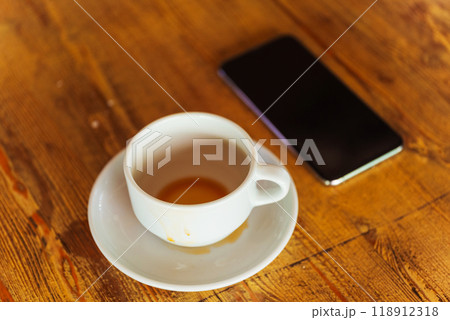 A coffee cup on a saucer beside a cell phone on a table 118912318