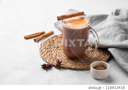 Glass mug with hot chocolate and milk foam on a light background with cinnamon sticks, anise star 118912418