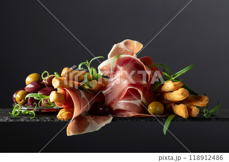 Prosciutto with bread sticks, olives, and rosemary on a black background. 118912486