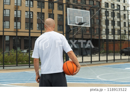 Basketball player with orange basketball ball on sports court courtyard. Basketball Hoop. View from back. Urban public park outside. Basketball player with orange basketball ball on sports court courtyard. Basketball Hoop. View from back. Urban public park outside. 118913522