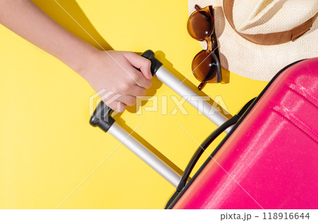 A hand holds a pink suitcase with travel accessories on a yellow background. A hand holds a pink suitcase with travel accessories on a yellow background. 118916644