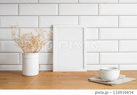 A frame with a blank canvas against a white tile wall and on a wooden tabletop with vase and cup. A frame with a blank canvas against a white tile wall and on a wooden tabletop with vase and cup. 118916654
