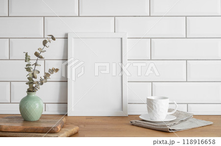A frame with a blank canvas against a white tile wall and on a wooden tabletop with vase A frame with a blank canvas against a white tile wall and on a wooden tabletop with vase 118916658