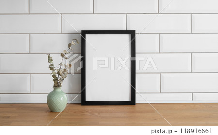 A frame with a blank canvas against a white tile wall and on a wooden tabletop with vase A frame with a blank canvas against a white tile wall and on a wooden tabletop with vase 118916661