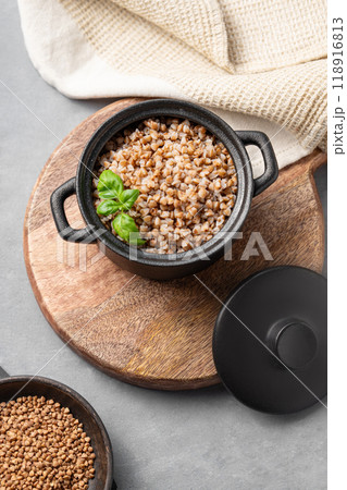 Buckwheat porridge in a black pan on a cutting board on a light background with raw grains in a bowl 118916813