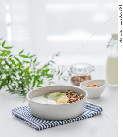 Granola with banana and kiwi on a white table with a green branch against the background of a window 118916861