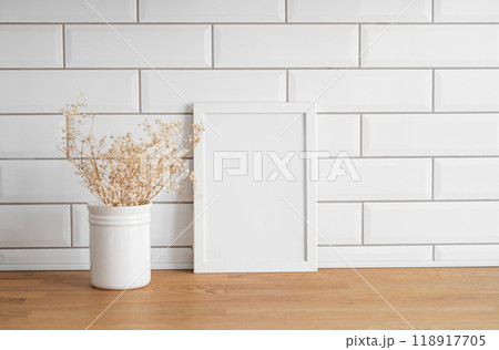 A frame with a blank canvas against a white tile wall and on a wooden tabletop with vase. A frame with a blank canvas against a white tile wall and on a wooden tabletop with vase. 118917705