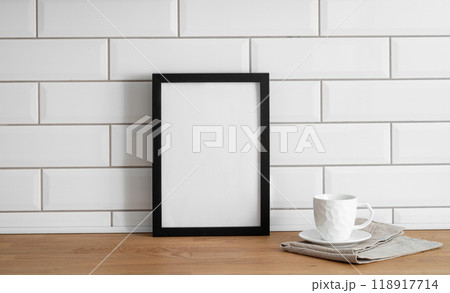 A frame with a blank canvas against a white tile wall and on a wooden tabletop with cup of tea. A frame with a blank canvas against a white tile wall and on a wooden tabletop with cup of tea. 118917714
