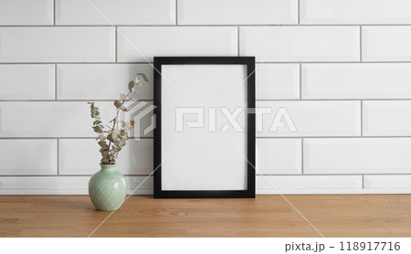 A  frame with a blank canvas against a white tile wall and on a wooden tabletop with vase 118917716