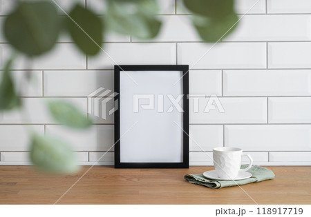 A black frame with a blank canvas against a white tile wall and on a wooden tabletop 118917719