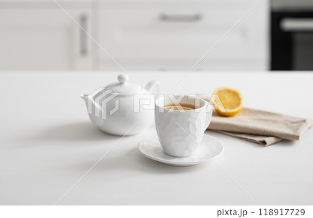 A cup of tea with lemon and a teapot on a white table against the background of a white kitchen. A cup of tea with lemon and a teapot on a white table against the background of a white kitchen. 118917729