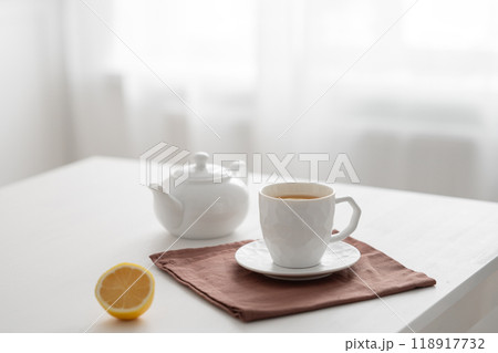 A cup of tea with lemon and a teapot on a white table against the background of a kitchen window. A cup of tea with lemon and a teapot on a white table against the background of a kitchen window. 118917732