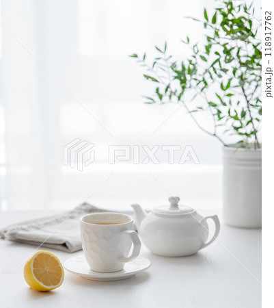 A cup of tea with lemon, a teapot and green branch on a white table against the background 118917762
