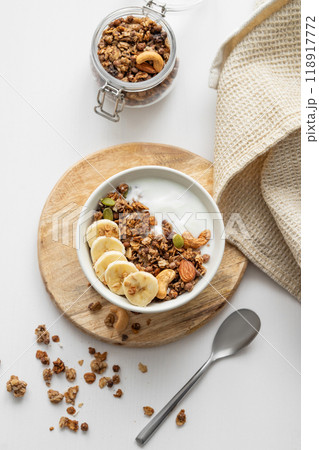 Granola with banana and nuts on a wooden board on a white table with a napkin and spoon. Granola with banana and nuts on a wooden board on a white table with a napkin and spoon. 118917772