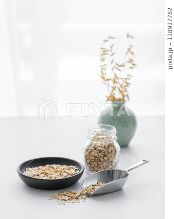 Oatmeal in a plate, scoop and in a jar on a white textured table in front of the kitchen window. Oatmeal in a plate, scoop and in a jar on a white textured table in front of the kitchen window. 118917782