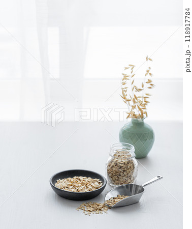 Oatmeal in a plate, scoop and in a jar on a white textured table in front of the kitchen window. 118917784