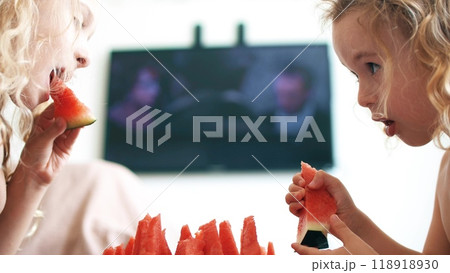 Two little girls eat fresh red watermelon slices and watch TV at home 118918930