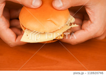 Home delivery. Man holds burger with hands. Hands holding homemade beef burger on wooden table. Ready to eat tasty fast food hamburger. Unhealthy food concept. Close up shot of young man eats burger. Home delivery. Man holds burger with hands. Hands holding homemade beef burger on wooden table. Ready to eat tasty fast food hamburger. Unhealthy food concept. Close up shot of young man eats burger. 118920746
