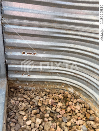 A detailed view of a corrugated metal window well with signs of rust and small holes. Gravel is scattered at the base, indicating outdoor exposure and wear over time.  118923930