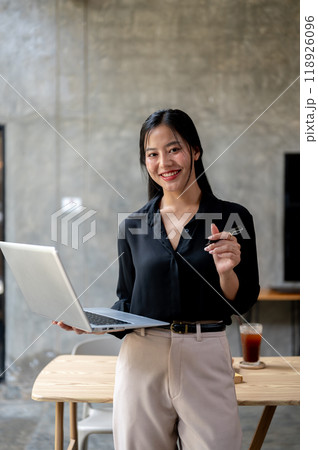 A confident Asian businesswoman is standing in her modern loft office with a laptop in her hand. 118926096