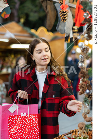 Cheerful girl in plaid coat choosing handmade Christmas decorations at fair Cheerful girl in plaid coat choosing handmade Christmas decorations at fair 118927301