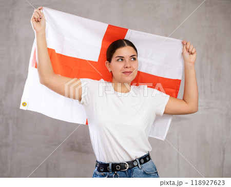Excited young woman in casual wear holding flag of England against gray wall background 118927623