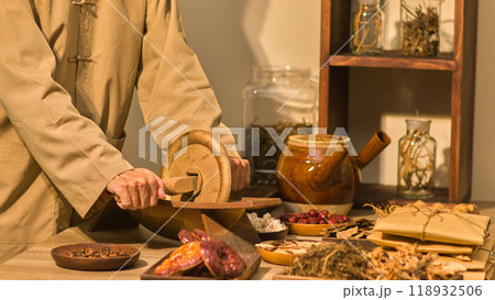 In a traditional eastern medicine room, herbs , packages, and other tools used in the field are arranged next to a herbalist who is grinding herbs with a wooden mill on a table. In a traditional eastern medicine room, herbs , packages, and other tools used in the field are arranged next to a herbalist who is grinding herbs with a wooden mill on a table. 118932506
