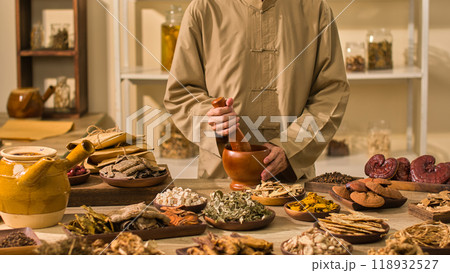 In a laboratory setting for a traditional medicine photo, a doctor dressed in brown grinds dry herbs with a wooden mortar and pestle while other herbal materials surround a table. 118932527