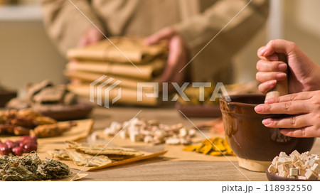 A herbalist is pounding angelica sinensis in the Oriental medication setting with a table full of herbs as another herbalist in beige clothing organizes the packets of wrapped herbal medication. 118932550