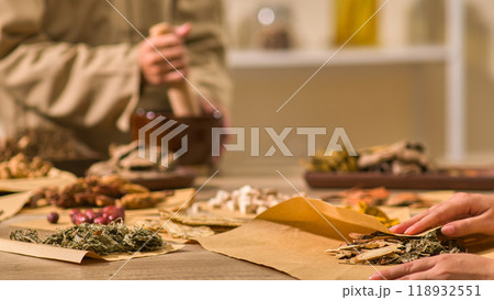One herbalist in traditional brown robes is pounding dried herbs, while another picks up a package of dried herbs at a traditional Chinese medicine on a table full of different herbs. 118932551