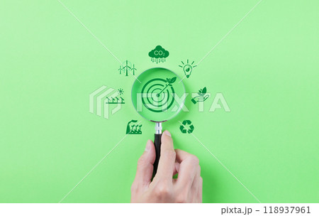Green Energy and Sustainability Concept. Hand holding a magnifying glass focusing on a target with a leaf, surrounded by green energy and sustainability icons on a green background. Carbon footprint, 118937961
