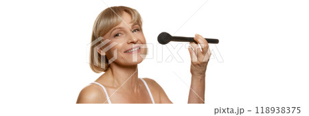 Banner. Smiling woman with makeup brush, about to apply powder, showing joy in her beauty routine against white studio background. Banner. Smiling woman with makeup brush, about to apply powder, showing joy in her beauty routine against white studio background. 118938375